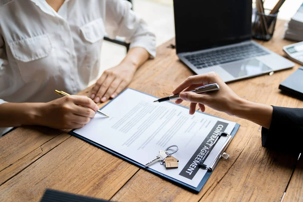 Crop a photo of a real estate agent helping clients read and sign contract documents at their desks with house designs. along with home insurance