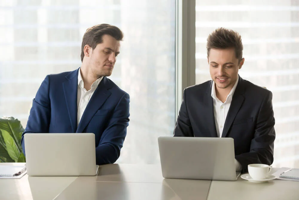 Curious businessman secretly looking at laptop screen of colleague