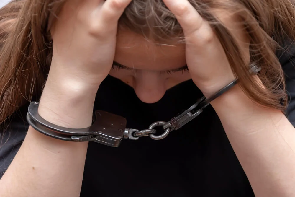 A young girl handcuffed hides her face on a gray background, close-up. Juvenile delinquent in a black T-shirt, criminal liability of minors.