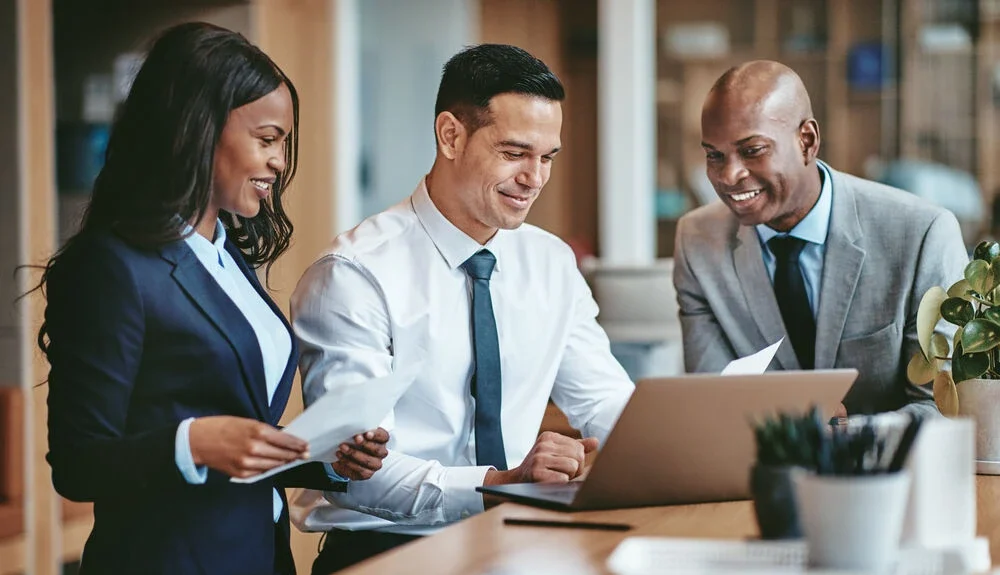 Smiling group of diverse businesspeople going over paperwork together and working on a laptop at a table in an office